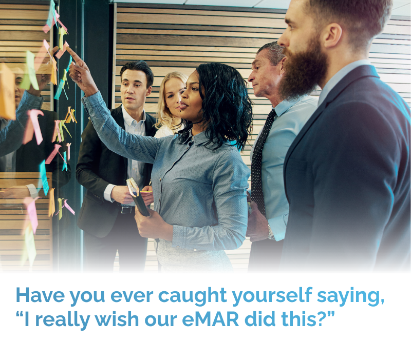 Several business people are gathered in an office, watching a woman point to a colorful sticky note on a glass partition covered with many notes.